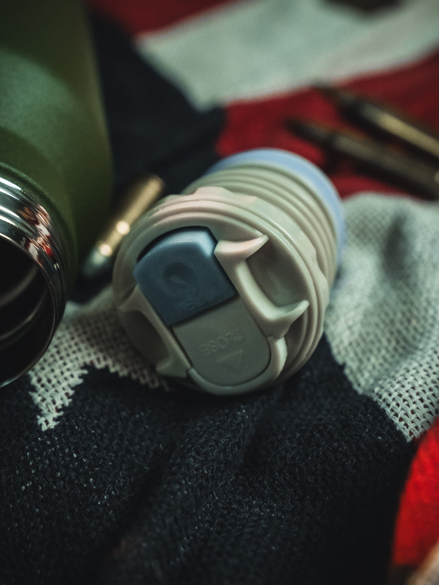 A close-up of a reusable bottle lid rests on red, white, and dark knitted fabric. In the background, a green Born United Private Label Bullet Thermos and metal accessories from the 32oz set are visible.