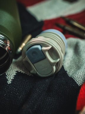 A close-up of a reusable bottle lid rests on red, white, and dark knitted fabric. In the background, a green Born United Private Label Bullet Thermos and metal accessories from the 32oz set are visible.