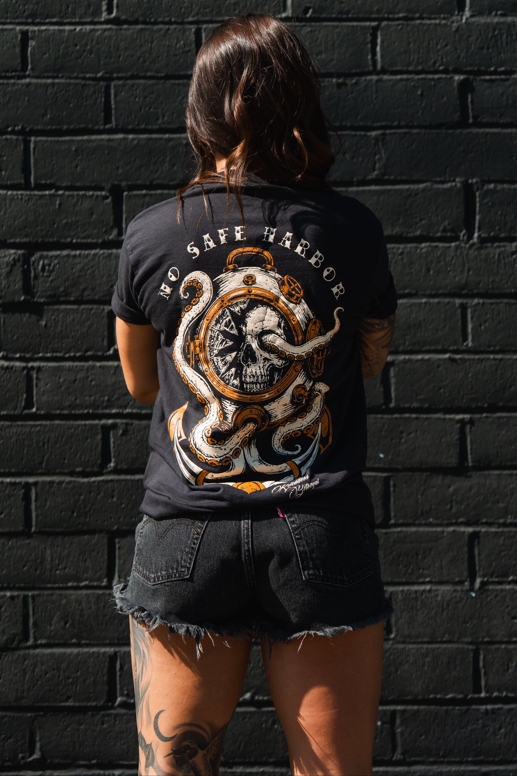 A woman with tattooed legs faces a black brick wall, wearing black shorts and the Relentless Betrayal No Safe Harbor Premium T-Shirt with an octopus skull design.