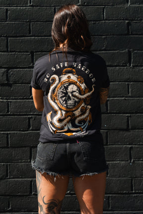A woman with tattooed legs faces a black brick wall, wearing black shorts and the Relentless Betrayal No Safe Harbor Premium T-Shirt with an octopus skull design.