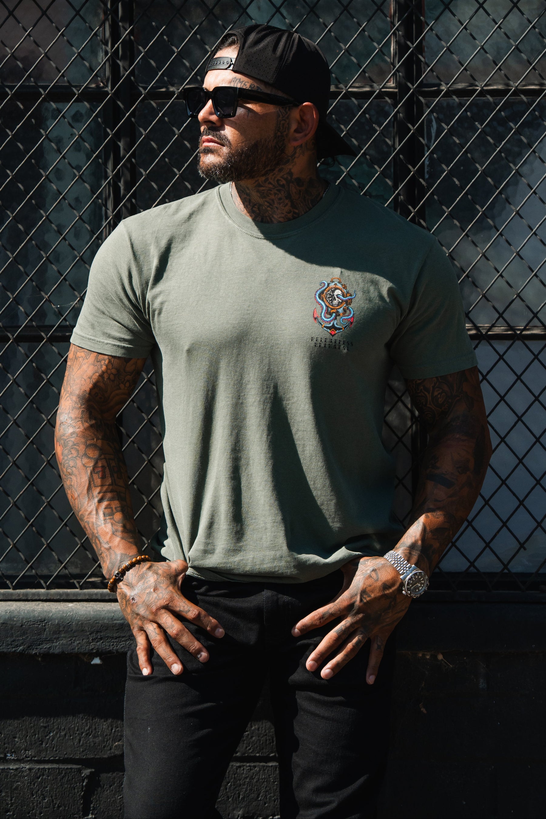 A tattooed man in a backward cap and Relentless Betrayal No Safe Harbor Premium T-Shirt - Olive stands by a metal fence, hands on hips, wearing sunglasses.