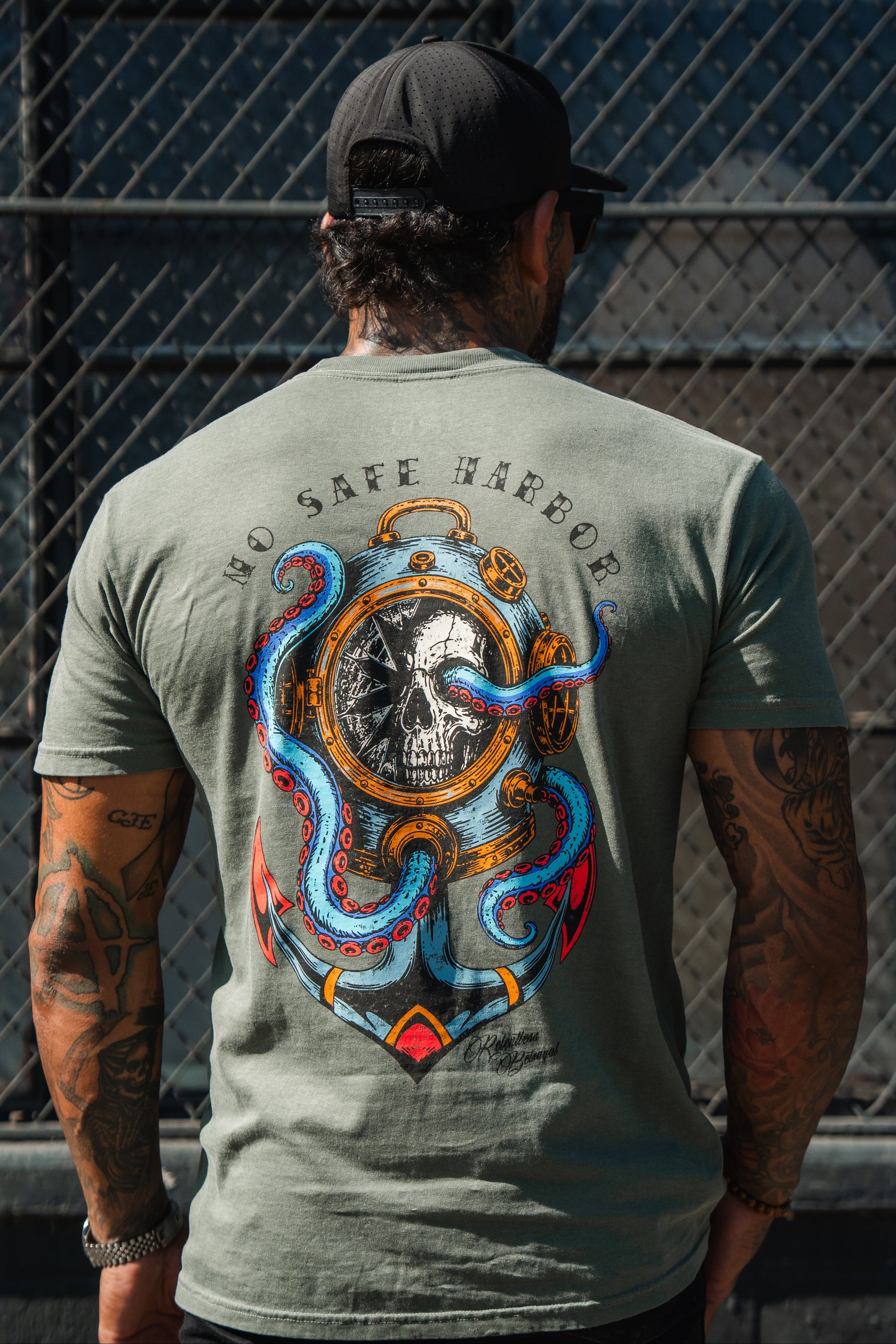 A man in a black cap wears the Relentless Betrayal No Safe Harbor Premium T-Shirt - Olive with a skull and octopus, standing by a metal fence.