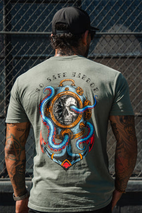 A man in a black cap wears the Relentless Betrayal No Safe Harbor Premium T-Shirt - Olive with a skull and octopus, standing by a metal fence.