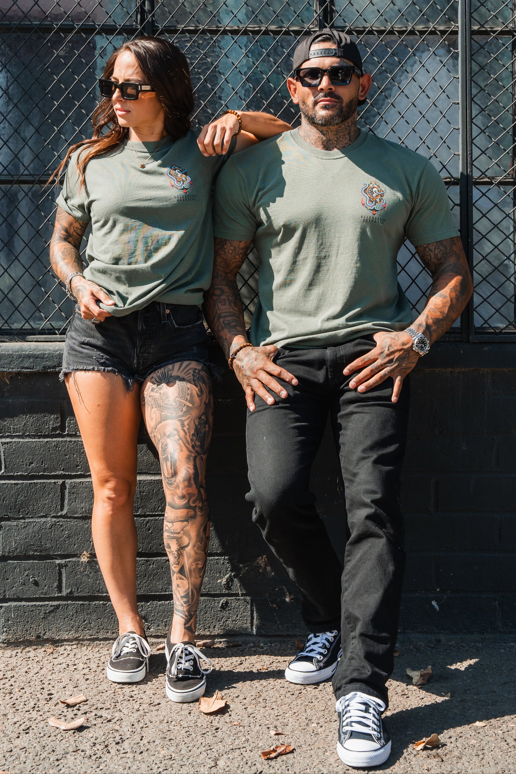 Two tattooed people in sunglasses and black sneakers wear matching Relentless Betrayal No Safe Harbor Premium T-Shirt - Olive, posing by a black brick wall and chain-link fence.