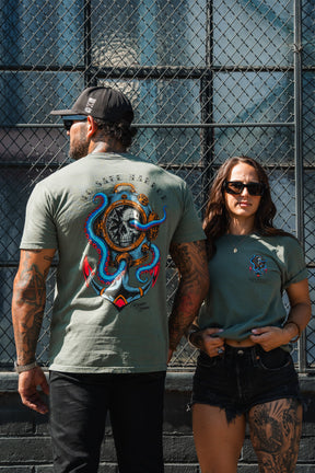 A man and woman in No Safe Harbor Premium T-Shirt - Olive by Relentless Betrayal pose by a chain-link fence, both in sunglasses and featuring bold Medusa designs.