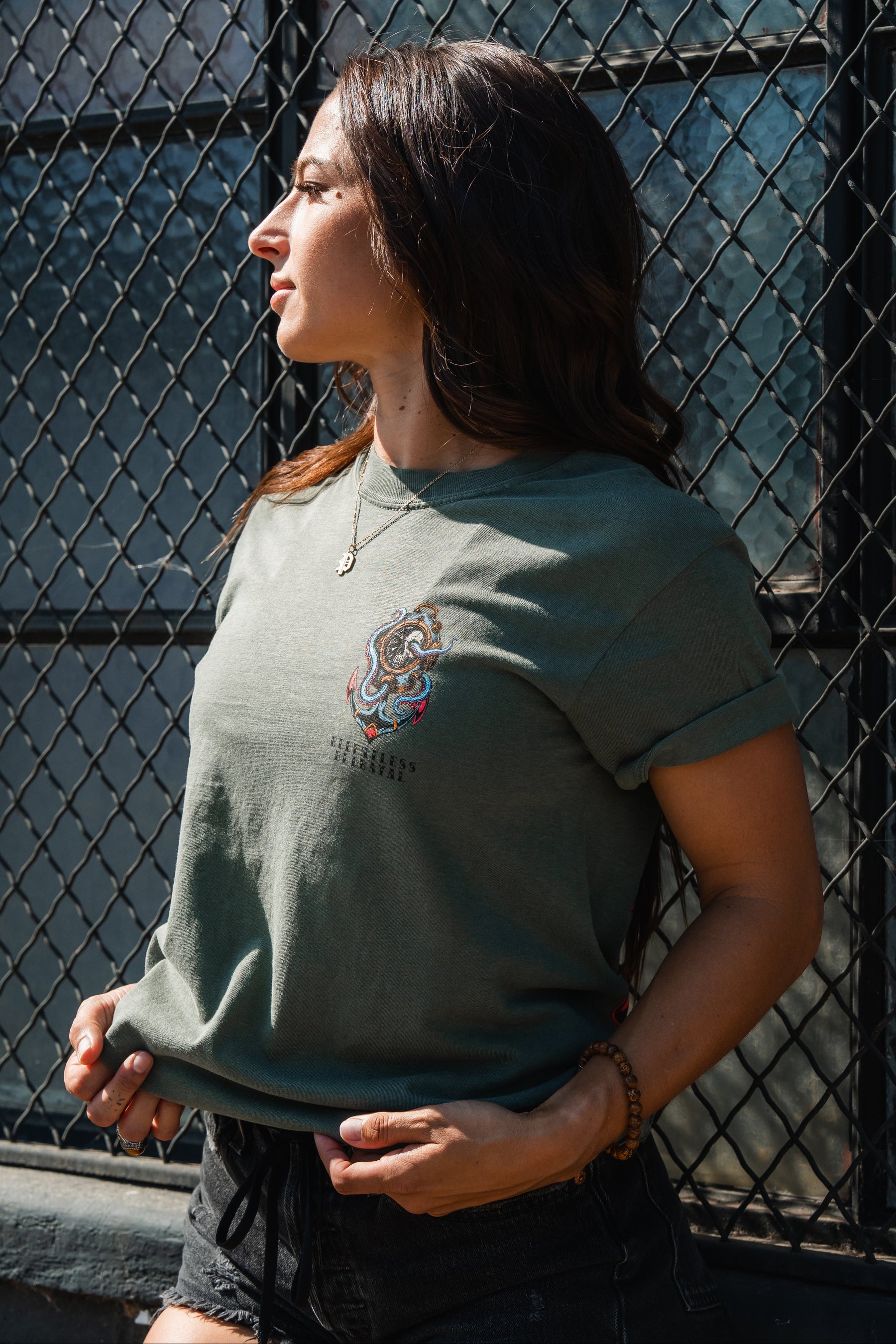 A woman in the Relentless Betrayal No Safe Harbor Premium T-Shirt - Olive and black shorts stands by a sunlit chain-link fence, looking left and holding her shirt.