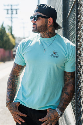 A tattooed man in the To The Ends Of The Earth Premium T-Shirt by Relentless Betrayal, sunglasses, and a backwards cap leans against a fence outdoors, looking to the side.