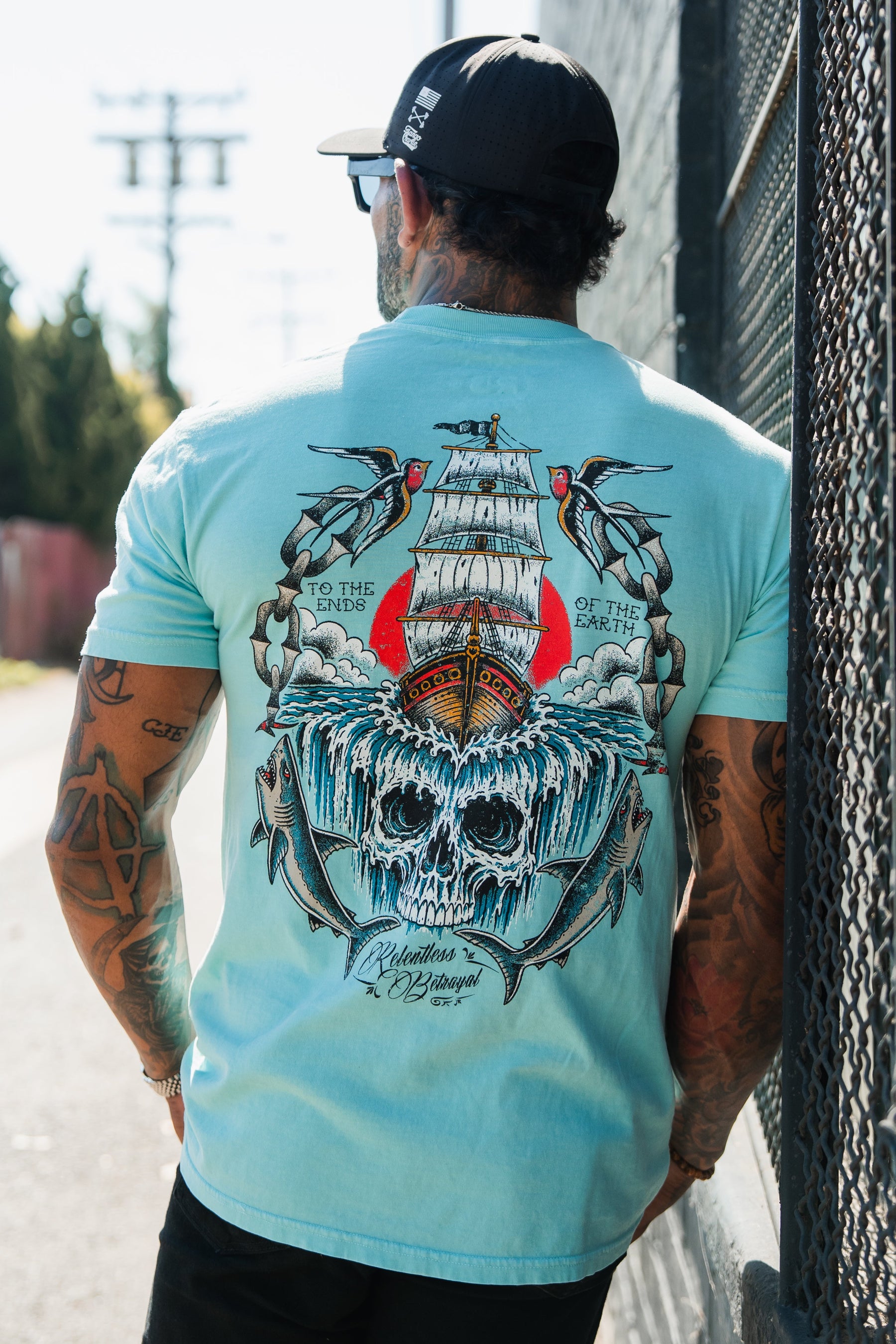 A man in a light blue To The Ends Of The Earth Premium T-Shirt by Relentless Betrayal stands outside near a fence, showing off the bold ship and skull graphic on the back.