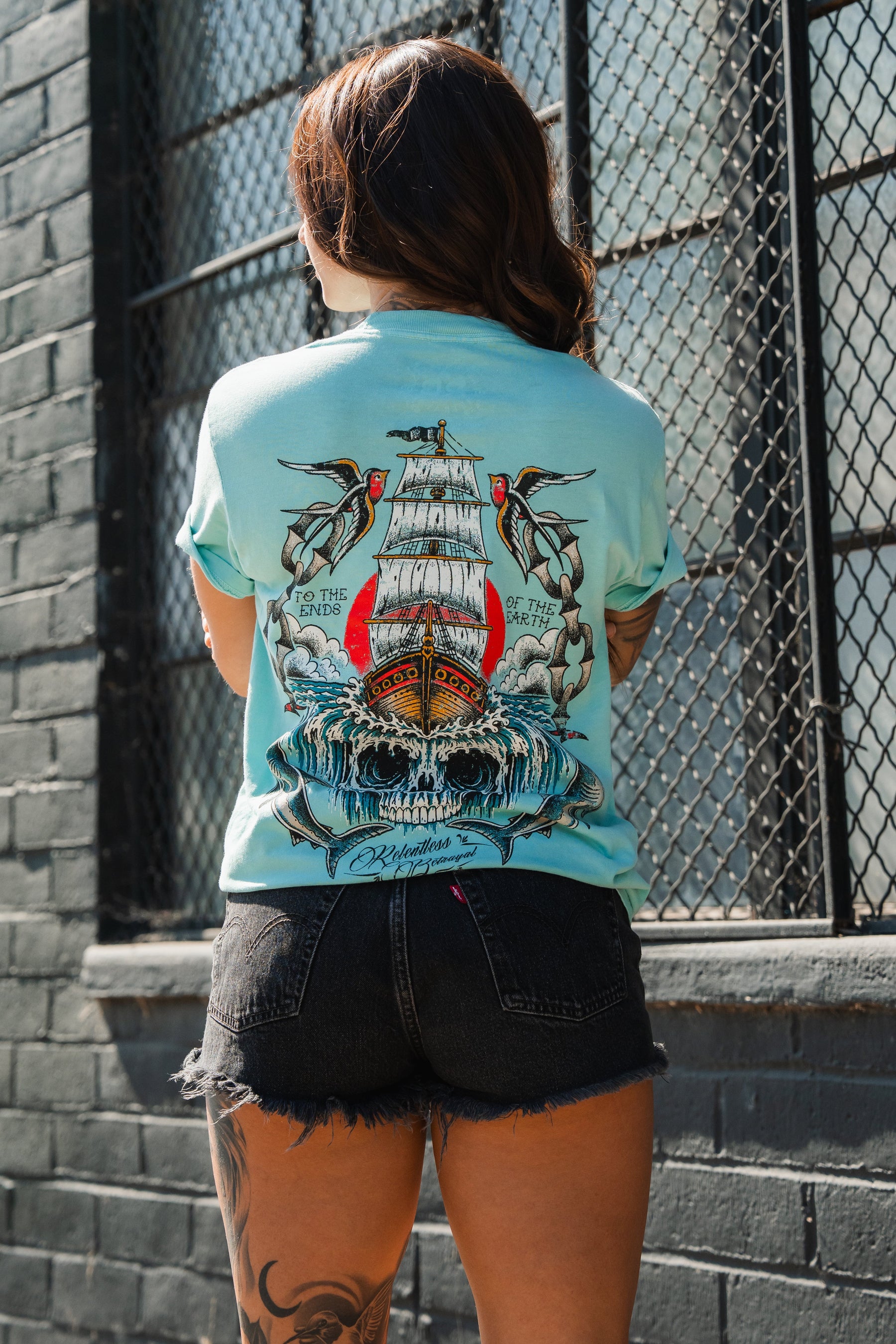 Person stands with back to camera in the Relentless Betrayal To The Ends Of The Earth Premium T-Shirt, black shorts, and a leg tattoo by a brick wall with metal-barred windows.