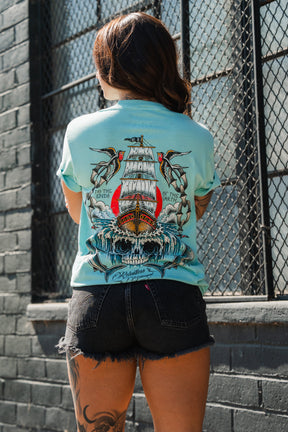 Person stands with back to camera in the Relentless Betrayal To The Ends Of The Earth Premium T-Shirt, black shorts, and a leg tattoo by a brick wall with metal-barred windows.