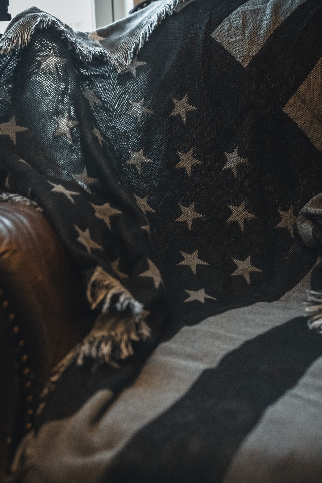 A close-up of the Born United Private Label Blackout American Flag Jacquard Blanket, featuring stars and stripes, draped over a brown leather chair with frayed edges and soft lighting highlighting its texture.