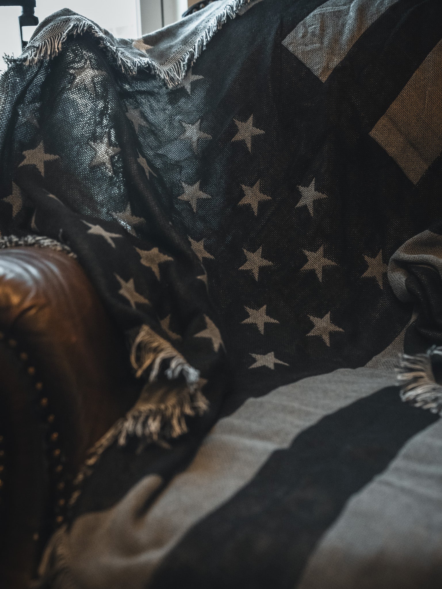 A close-up of the Born United Private Label Blackout American Flag Jacquard Blanket, featuring stars and stripes, draped over a brown leather chair with frayed edges and soft lighting highlighting its texture.
