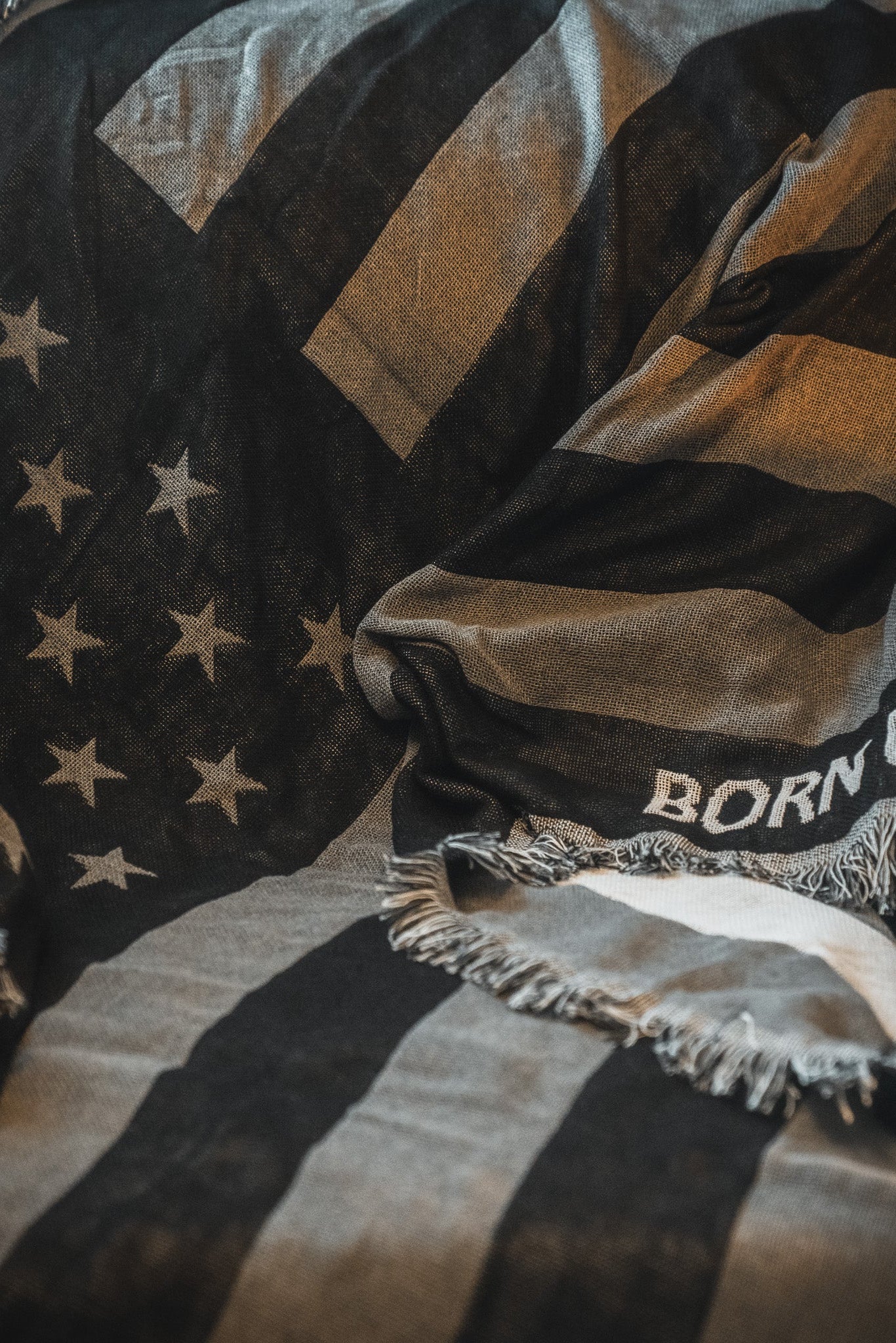 The Blackout American Flag Jacquard Blanket by Born United Private Label features a black and white flag design with fringed edges and the partial text "BORN UN" draped across a surface.
