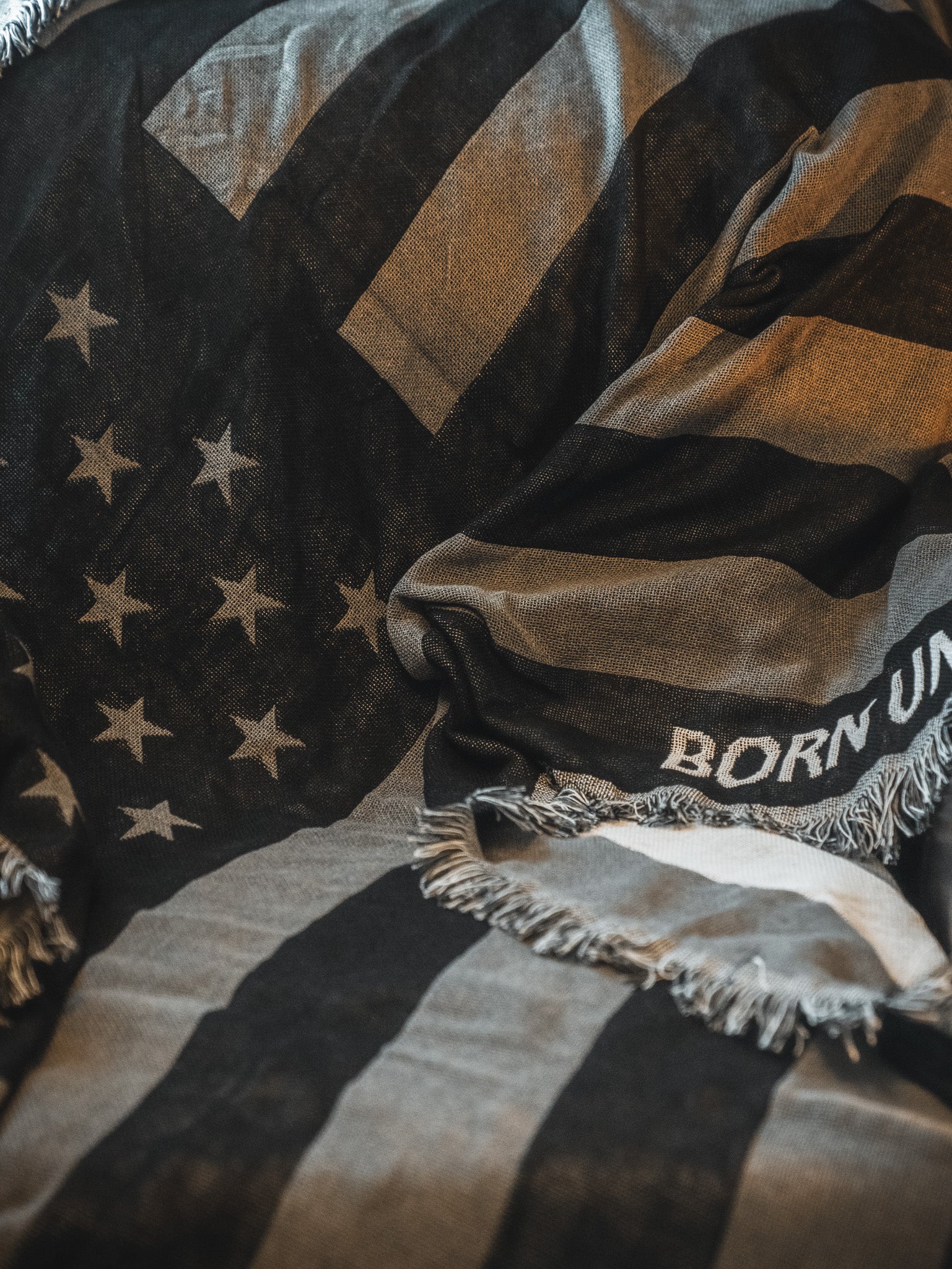 The Blackout American Flag Jacquard Blanket by Born United Private Label features a black and white flag design with fringed edges and the partial text "BORN UN" draped across a surface.
