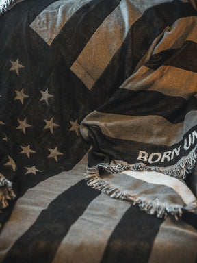 The Blackout American Flag Jacquard Blanket by Born United Private Label features a black and white flag design with fringed edges and the partial text "BORN UN" draped across a surface.