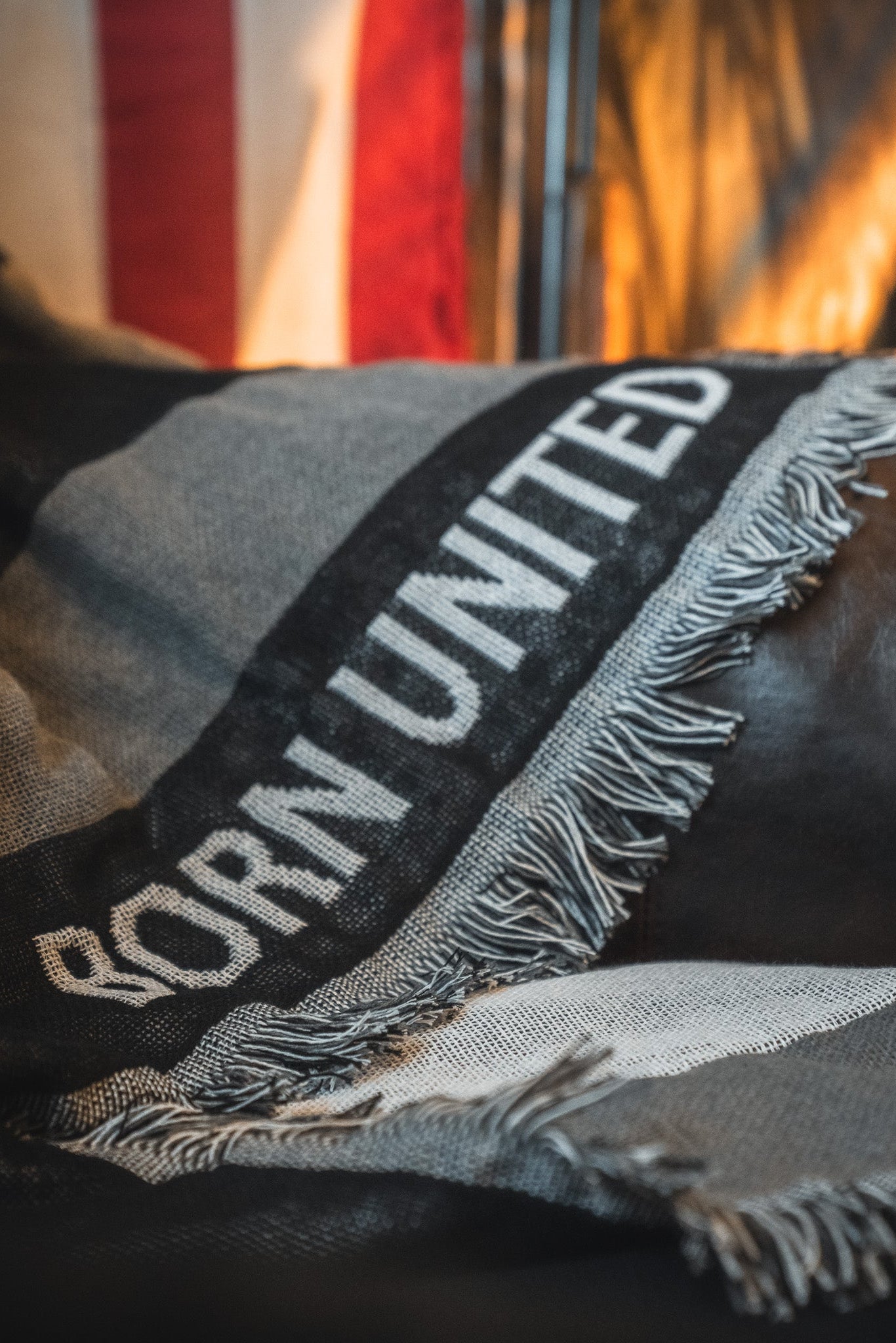 A close-up of the Born United Private Label Blackout American Flag Jacquard Blanket shows "BORN UNITED" woven in white on black and gray fabric, with a fringed edge. A blurred American flag appears in the background.