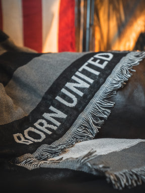 A close-up of the Born United Private Label Blackout American Flag Jacquard Blanket shows "BORN UNITED" woven in white on black and gray fabric, with a fringed edge. A blurred American flag appears in the background.