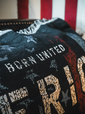 A close-up of the American Heroes Jacquard Blanket by Born United Private Label, showcasing woven stars and stripes in red, white, and blue with the phrase "BORN UNITED." A blurred American flag appears in the background.