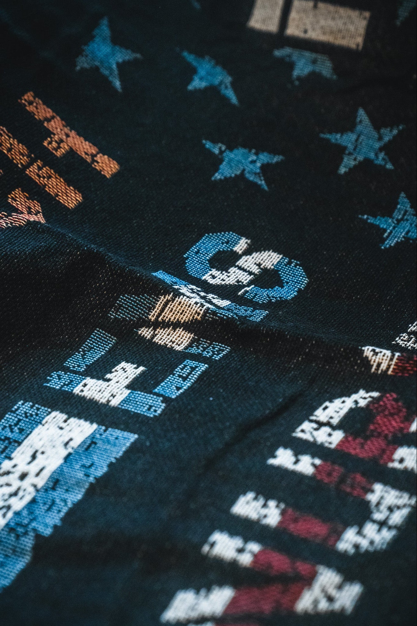 A close-up of the Born United Private Label American Heroes Jacquard Blanket, showing dark fabric with vibrant abstract patterns, numbers, letters, and stars in blue, orange, white, and red, highlighting its rich texture and visible folds.