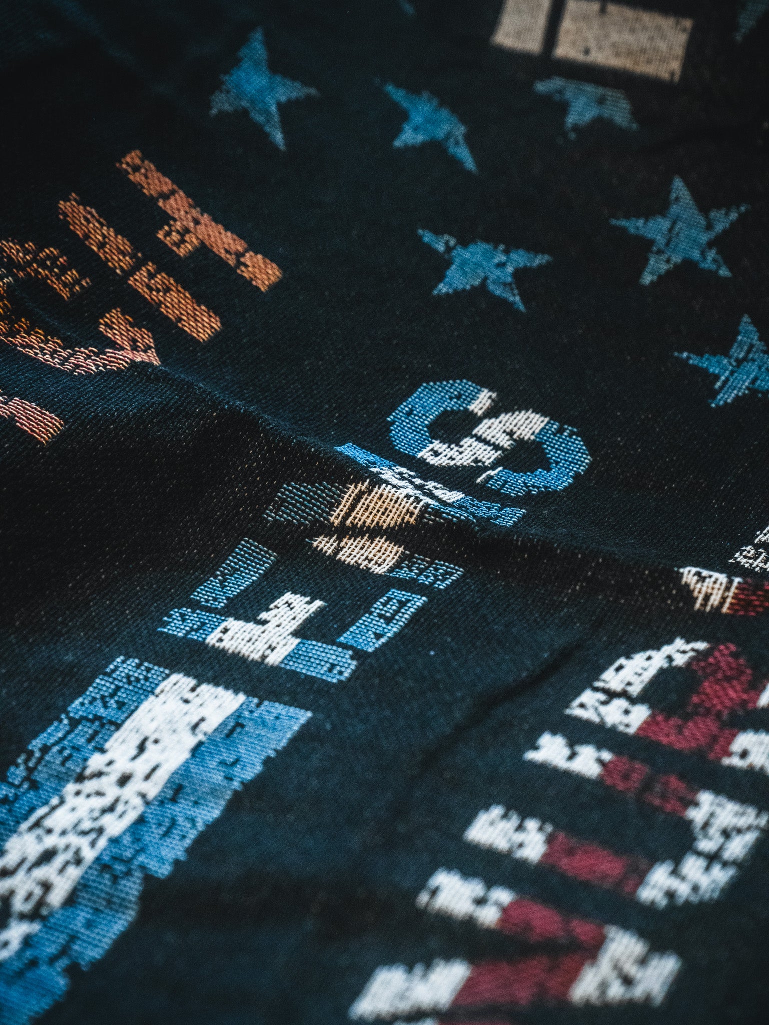 A close-up of the Born United Private Label American Heroes Jacquard Blanket, showing dark fabric with vibrant abstract patterns, numbers, letters, and stars in blue, orange, white, and red, highlighting its rich texture and visible folds.