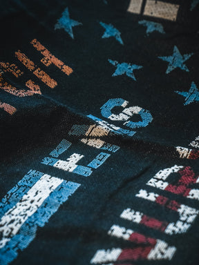 A close-up of the Born United Private Label American Heroes Jacquard Blanket, showing dark fabric with vibrant abstract patterns, numbers, letters, and stars in blue, orange, white, and red, highlighting its rich texture and visible folds.