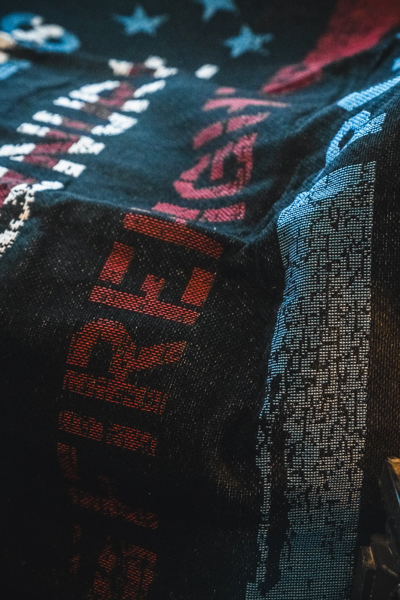 The American Heroes Jacquard Blanket by Born United Private Label features a textured black design with bold red and white lettering, blue and white stars, and abstract patterns for a patriotic look. Lighting accentuates its intricate weave and details.