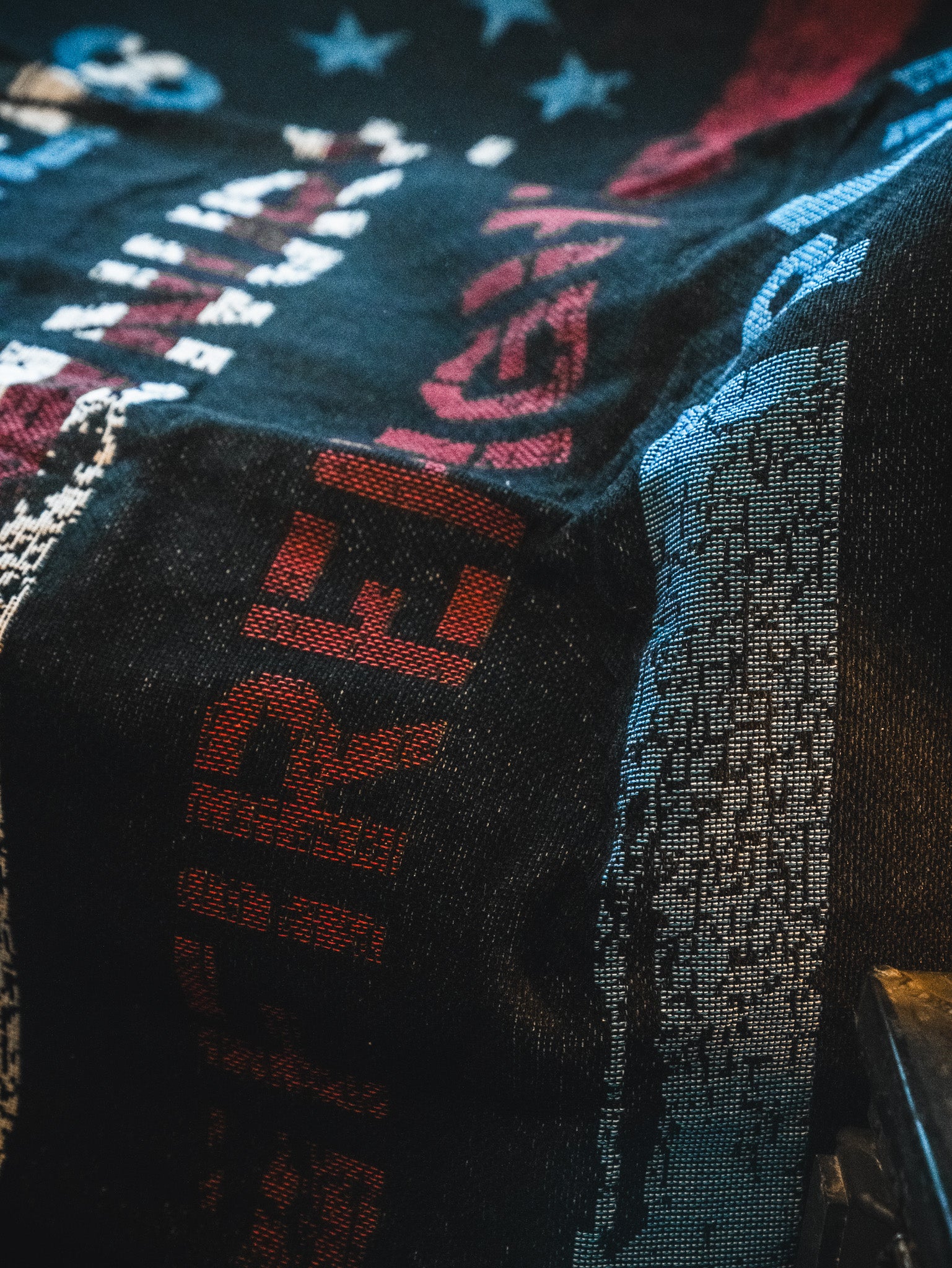 The American Heroes Jacquard Blanket by Born United Private Label features a textured black design with bold red and white lettering, blue and white stars, and abstract patterns for a patriotic look. Lighting accentuates its intricate weave and details.