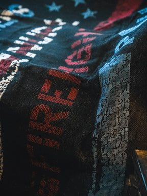 The American Heroes Jacquard Blanket by Born United Private Label features a textured black design with bold red and white lettering, blue and white stars, and abstract patterns for a patriotic look. Lighting accentuates its intricate weave and details.