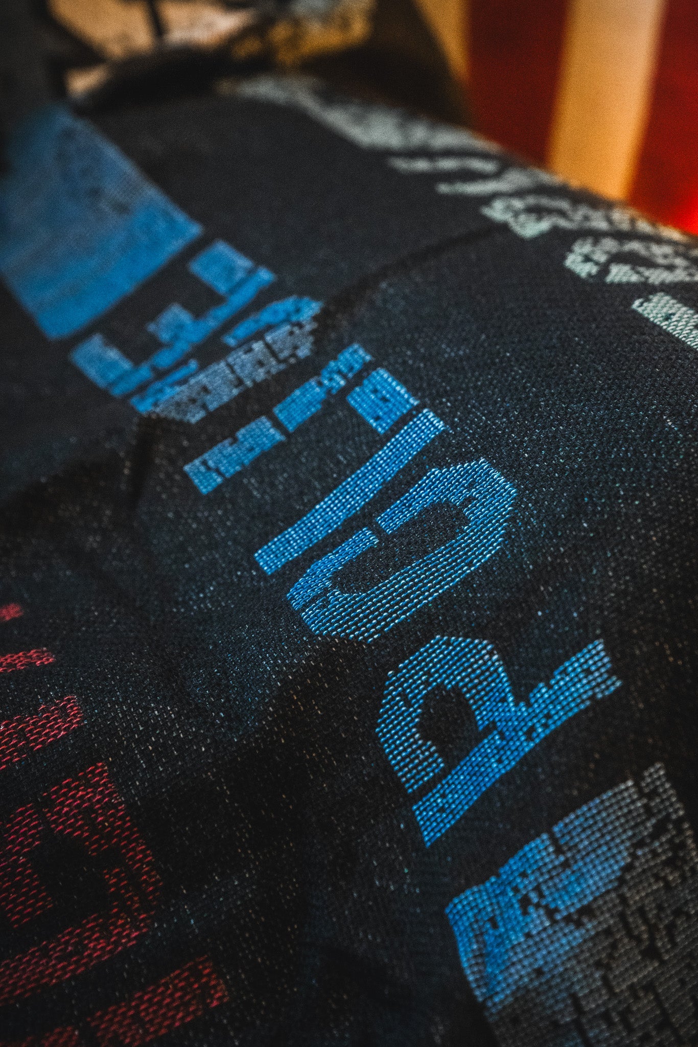 Close-up of the American Heroes Jacquard Blanket by Born United Private Label, featuring dark fabric with blue and red pixelated letters and numbers in a diagonal pattern, partially showing a blurred American flag in the background.