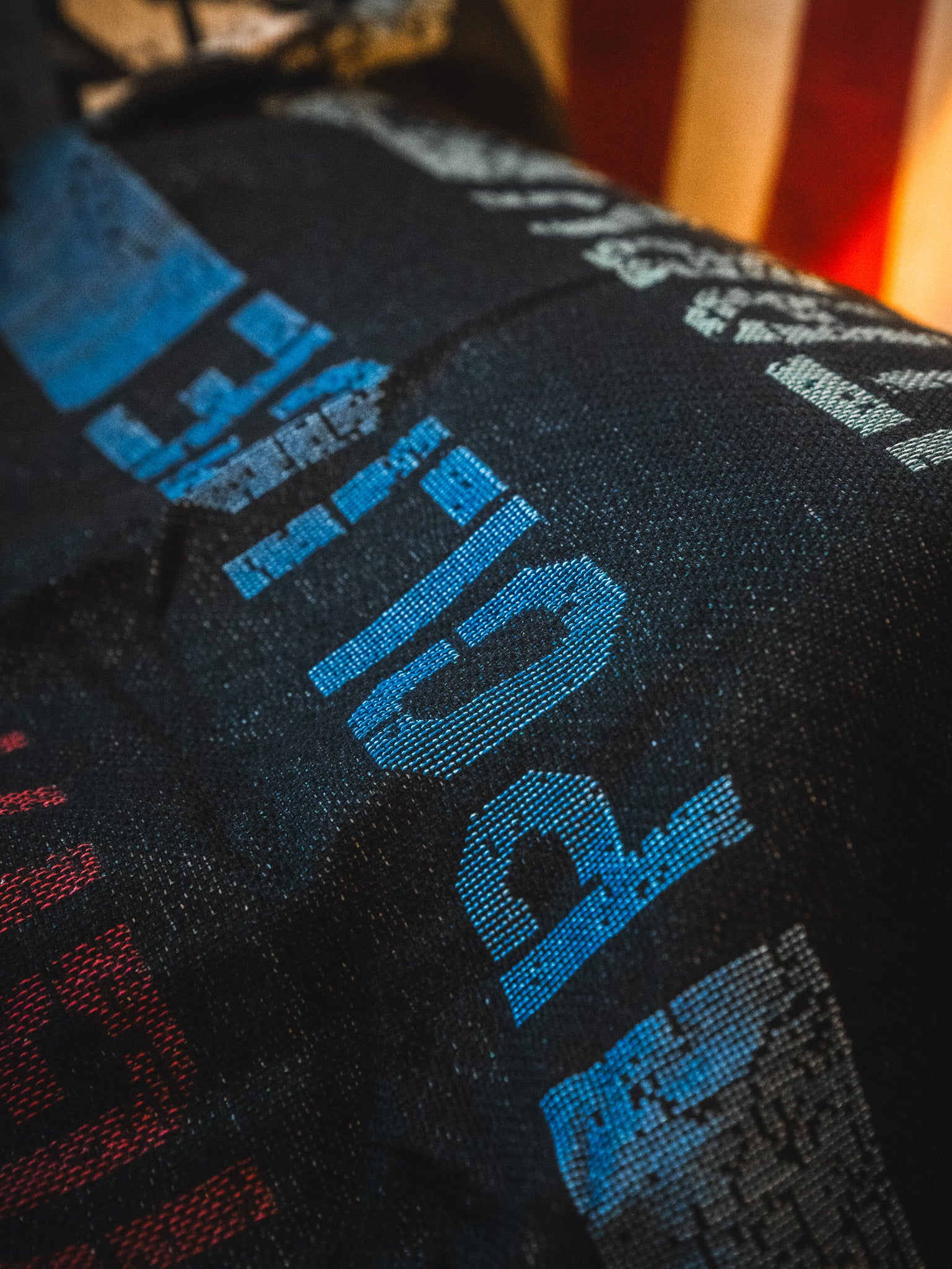 Close-up of the American Heroes Jacquard Blanket by Born United Private Label, featuring dark fabric with blue and red pixelated letters and numbers in a diagonal pattern, partially showing a blurred American flag in the background.