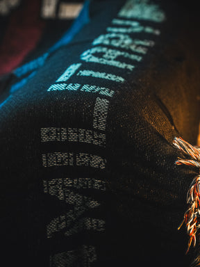 Close-up of dark denim fabric with light blue digital-style stitches and a decorative braided detail in orange, silver, and beige—echoing the design of the American Heroes Jacquard Blanket by Born United Private Label.