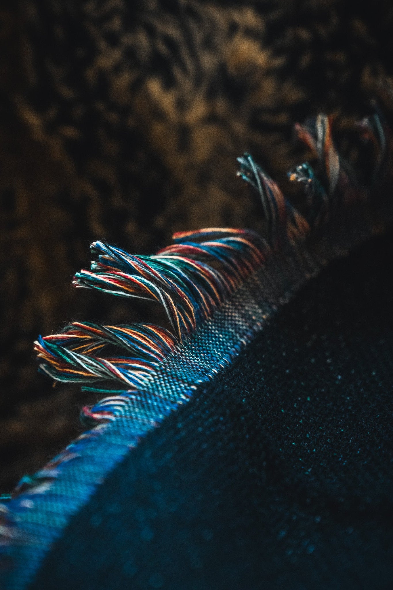 Close-up of multicolored, frayed fringe on the American Heroes Jacquard Blanket by Born United Private Label, set against a blurred brown and black background.