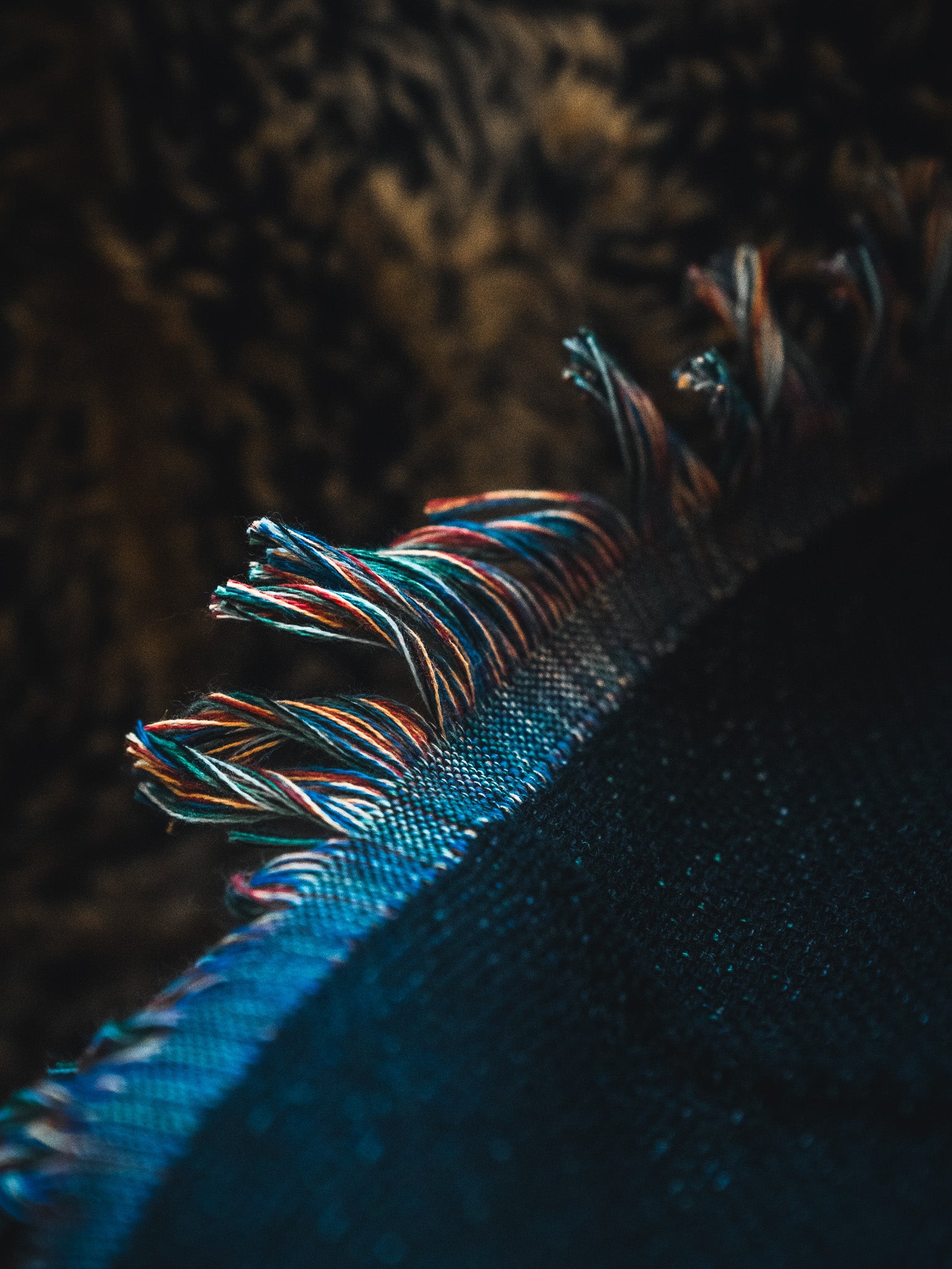 Close-up of multicolored, frayed fringe on the American Heroes Jacquard Blanket by Born United Private Label, set against a blurred brown and black background.