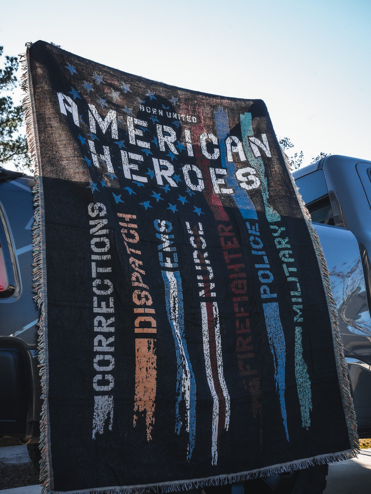 The Born United Private Label American Heroes Jacquard Blanket displays “American Heroes” with colorful faded stripes labeled Corrections, Dispatch, EMS, Firefighter, Police, and Military—perfect for draping over a vehicle.