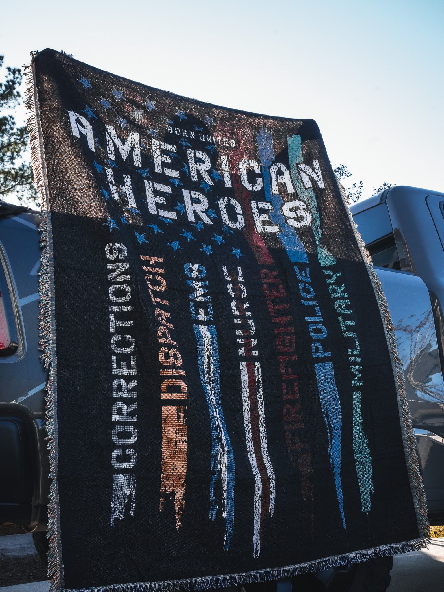 The Born United Private Label American Heroes Jacquard Blanket displays “American Heroes” with colorful faded stripes labeled Corrections, Dispatch, EMS, Firefighter, Police, and Military—perfect for draping over a vehicle.