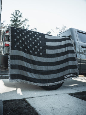 A Blackout American Flag Jacquard Blanket by Born United Private Label, featuring "BORN UNITED" in one corner, is draped over a pickup truck's tailgate parked outdoors on a sunny day.