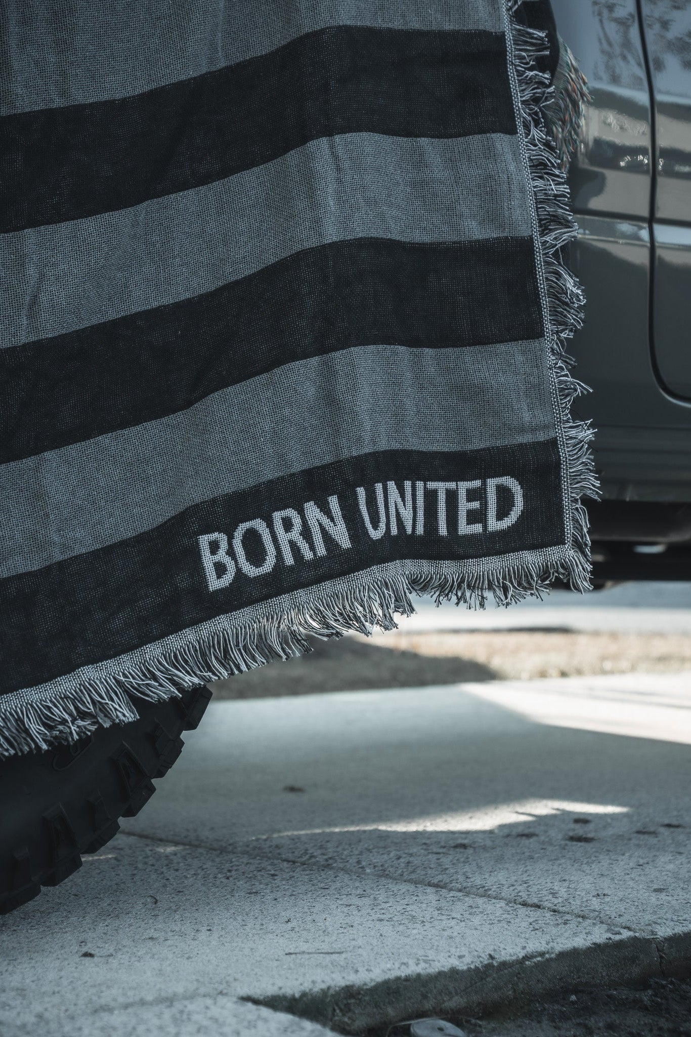A Blackout American Flag Jacquard Blanket from Born United Private Label, featuring gray and black stripes and fringed edges, is draped over the side of a vehicle, partially covering a tire on a sunlit street.