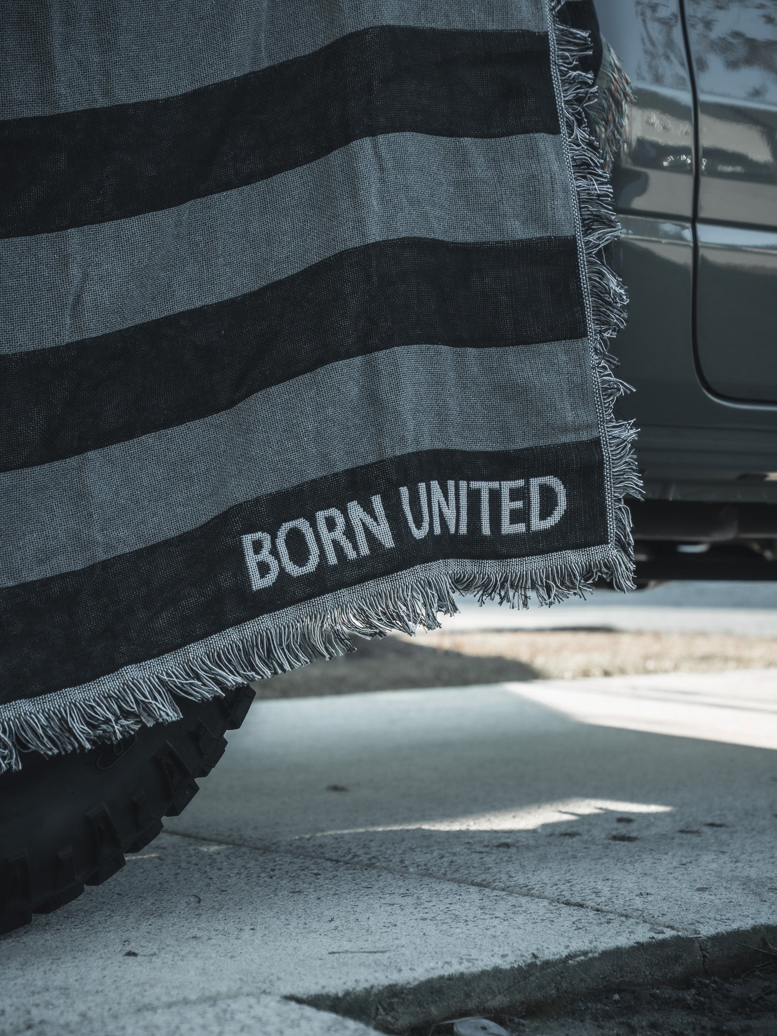 A Blackout American Flag Jacquard Blanket from Born United Private Label, featuring gray and black stripes and fringed edges, is draped over the side of a vehicle, partially covering a tire on a sunlit street.
