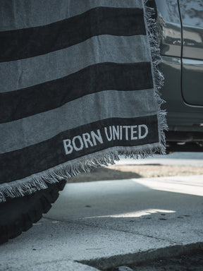A Blackout American Flag Jacquard Blanket from Born United Private Label, featuring gray and black stripes and fringed edges, is draped over the side of a vehicle, partially covering a tire on a sunlit street.