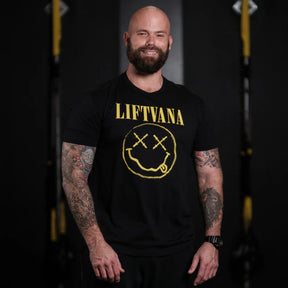 A smiling, bald, bearded man with tattooed arms stands by gym equipment, wearing the "Liftvana" Men's Tee from Tango Charlie Apparel—showcasing a black shirt with a yellow smiley face and X eyes for true lifter spirit and grunge style.