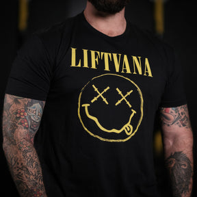 A muscular man with tattooed arms wears the Liftvana Mens Tee by Tango Charlie Apparel, featuring Lift•Va•Na text and a smiley face with barbell eyes—grunge vibes inspired by the iconic Nirvana logo.
