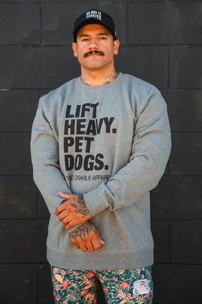 A man with a mustache stands against a black wall, wearing a black cap, colorful floral shorts, and the Lift Heavy. Pet Dogs Crewneck by Tango Charlie Apparel. He has hand tattoos and looks like someone who truly loves dogs.