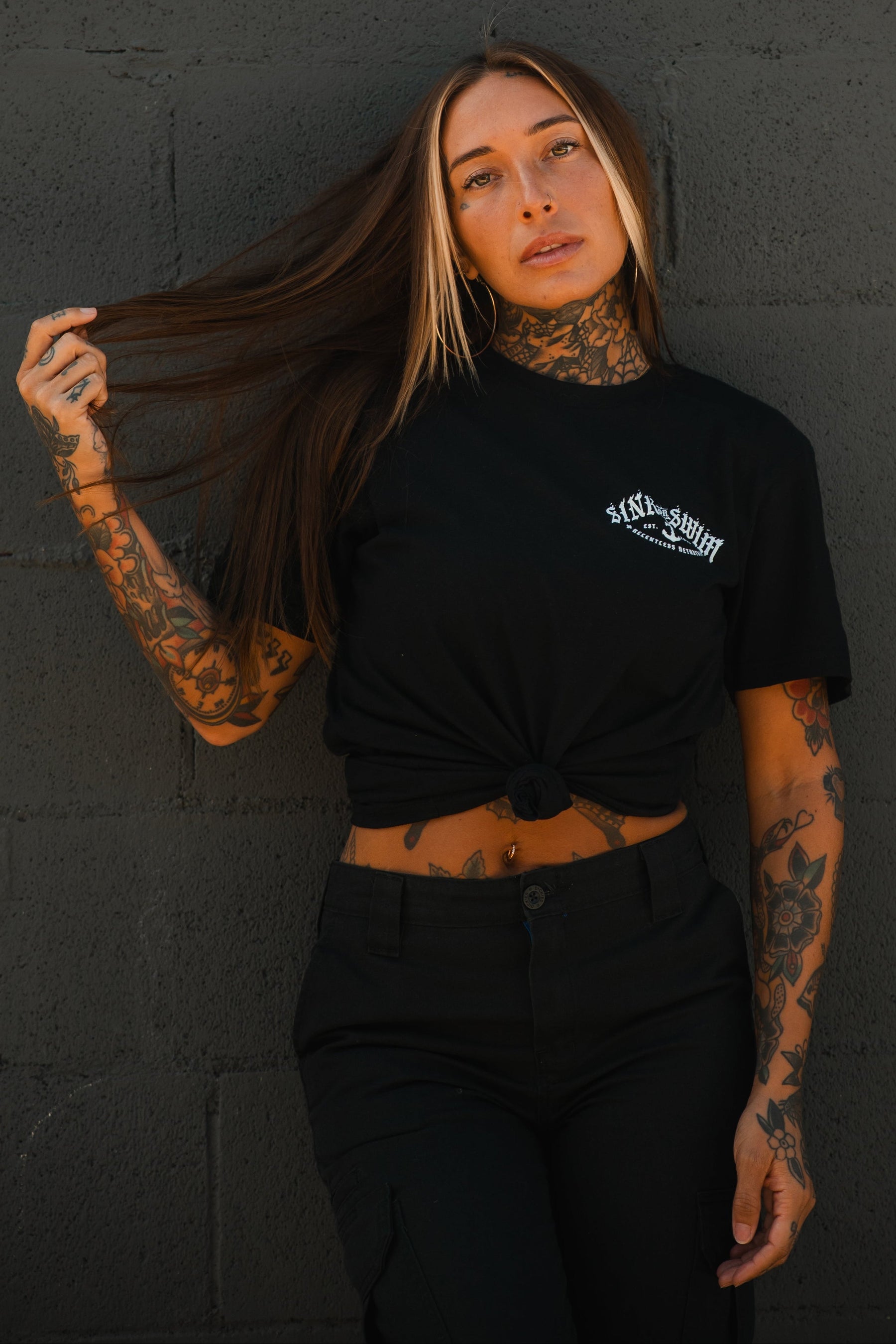 A woman with long brown hair, blonde highlights, and visible tattoos poses against a gray wall, holding her hair while wearing a black Relentless Betrayal Dark Waters SOS T-Shirt knotted at the waist with black pants.