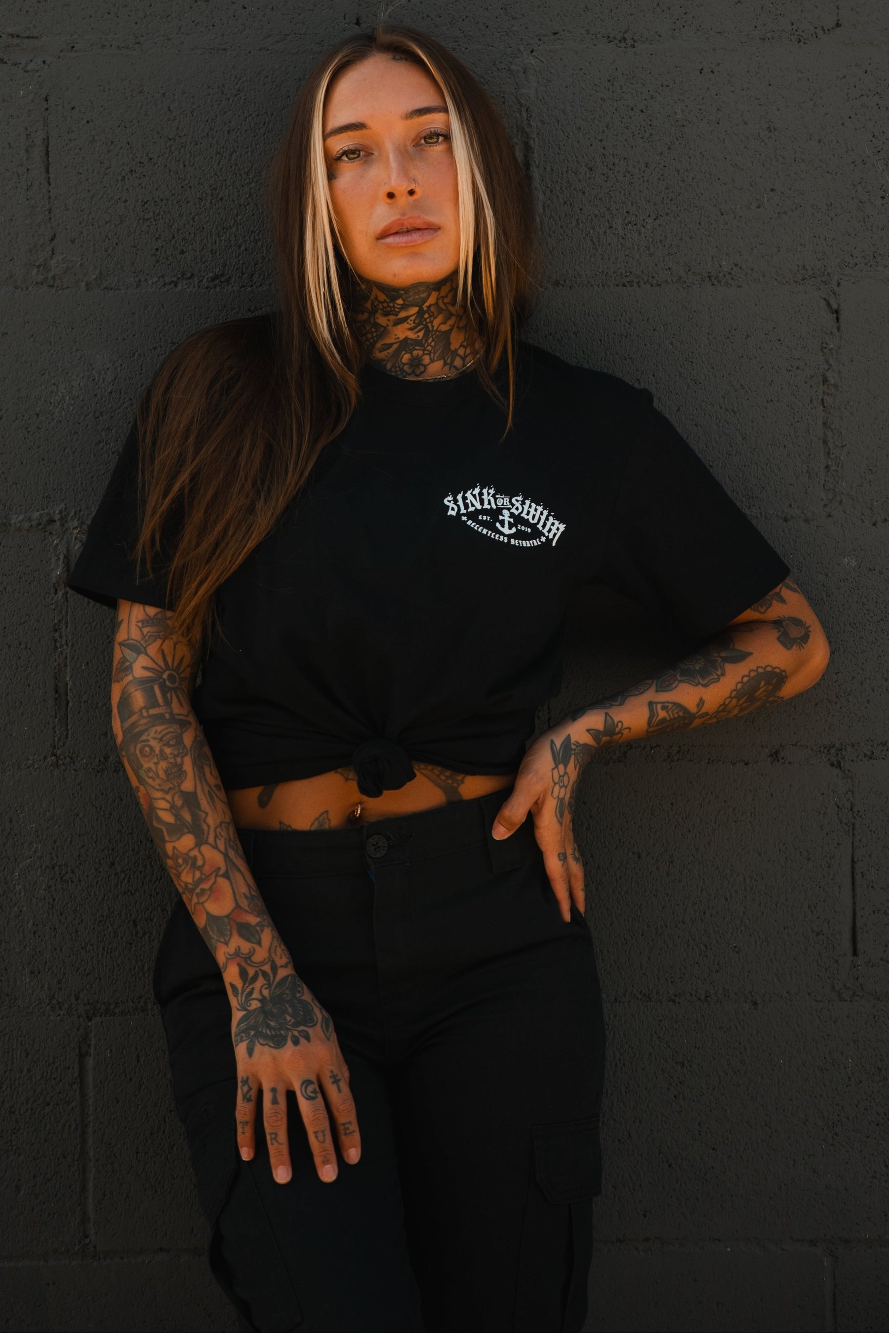 A woman with long brown hair and visible tattoos poses confidently against a dark gray brick wall, wearing the Relentless Betrayal Dark Waters SOS T-Shirt—an ultra-soft unisex tee—knotted at the waist with black pants.