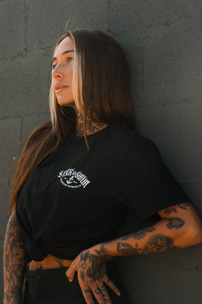 A woman with long brown hair and tattoos leans on a gray brick wall, wearing the Relentless Betrayal Dark Waters SOS T-Shirt—a black unisex cotton tee with a white graphic design—as she gazes calmly to the side.