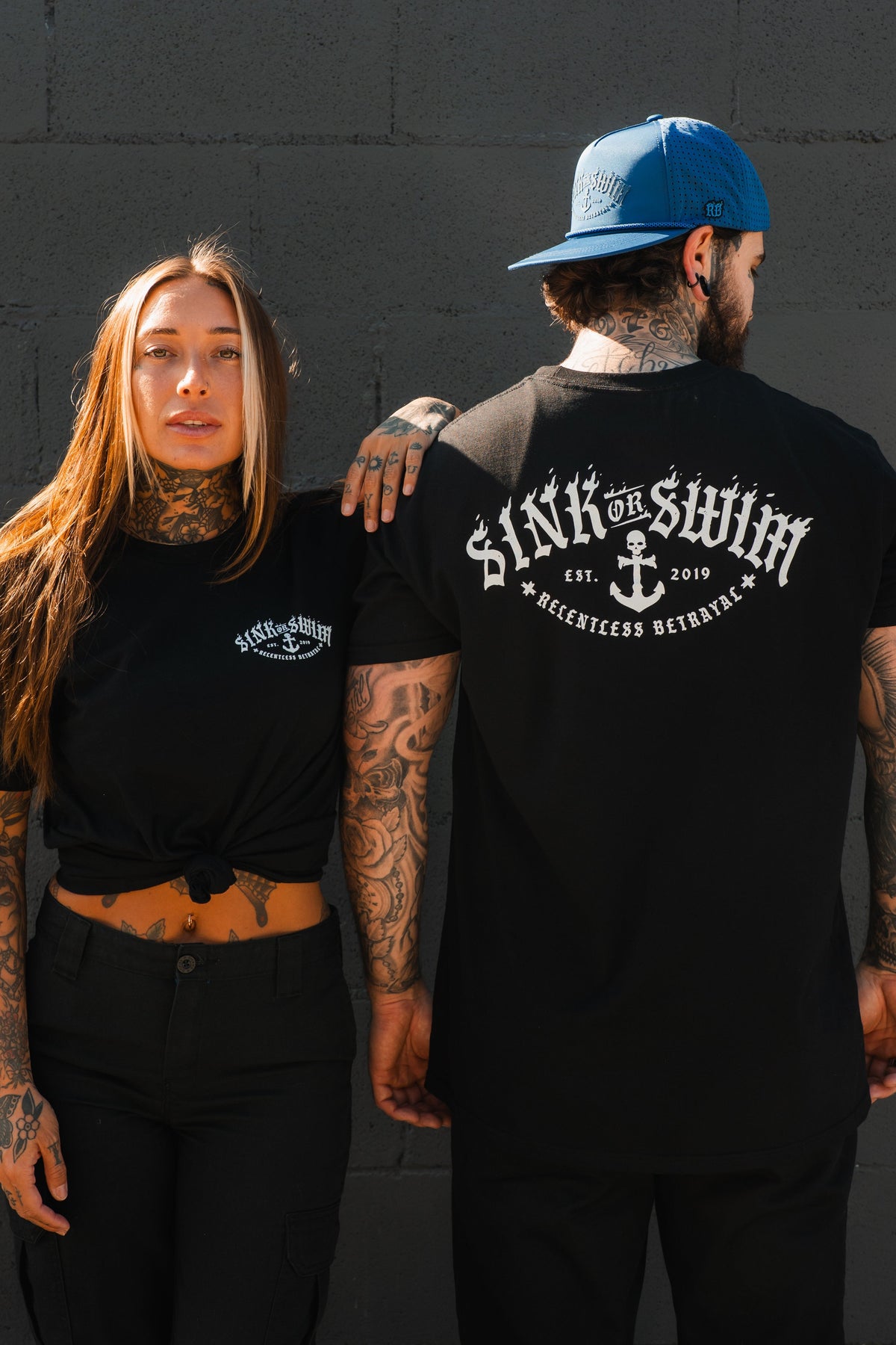 A tattooed woman and man wear black Relentless Betrayal Dark Waters SOS T-Shirts; she faces front, he shows the large anchor graphic and text on the back, standing against a gray brick wall.