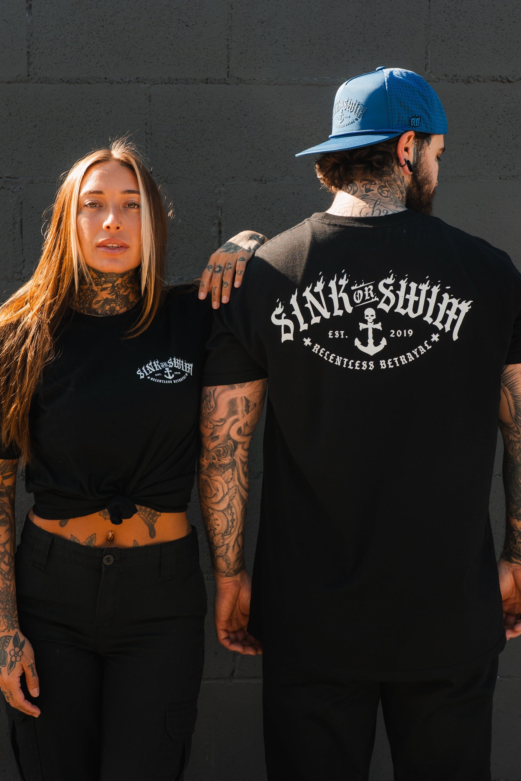 A tattooed woman and man wear black Relentless Betrayal Dark Waters SOS T-Shirts; she faces front, he shows the large anchor graphic and text on the back, standing against a gray brick wall.