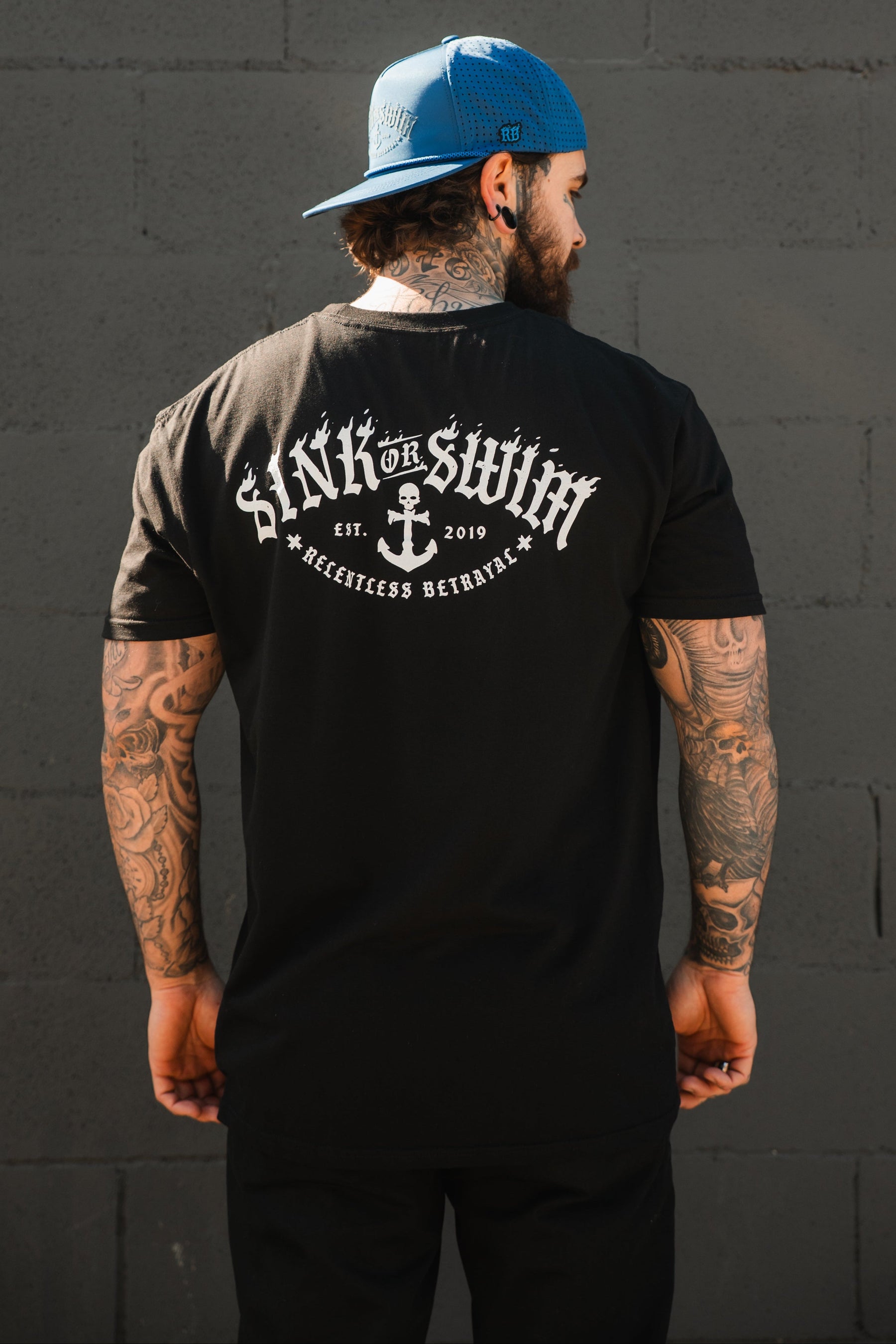 A tattooed man in a backwards blue cap and Relentless Betrayal's Dark Waters SOS T-Shirt, featuring "Sink or Swim" and an anchor graphic on the back, stands before a gray brick wall.