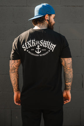 A tattooed man in a backwards blue cap and Relentless Betrayal's Dark Waters SOS T-Shirt, featuring "Sink or Swim" and an anchor graphic on the back, stands before a gray brick wall.