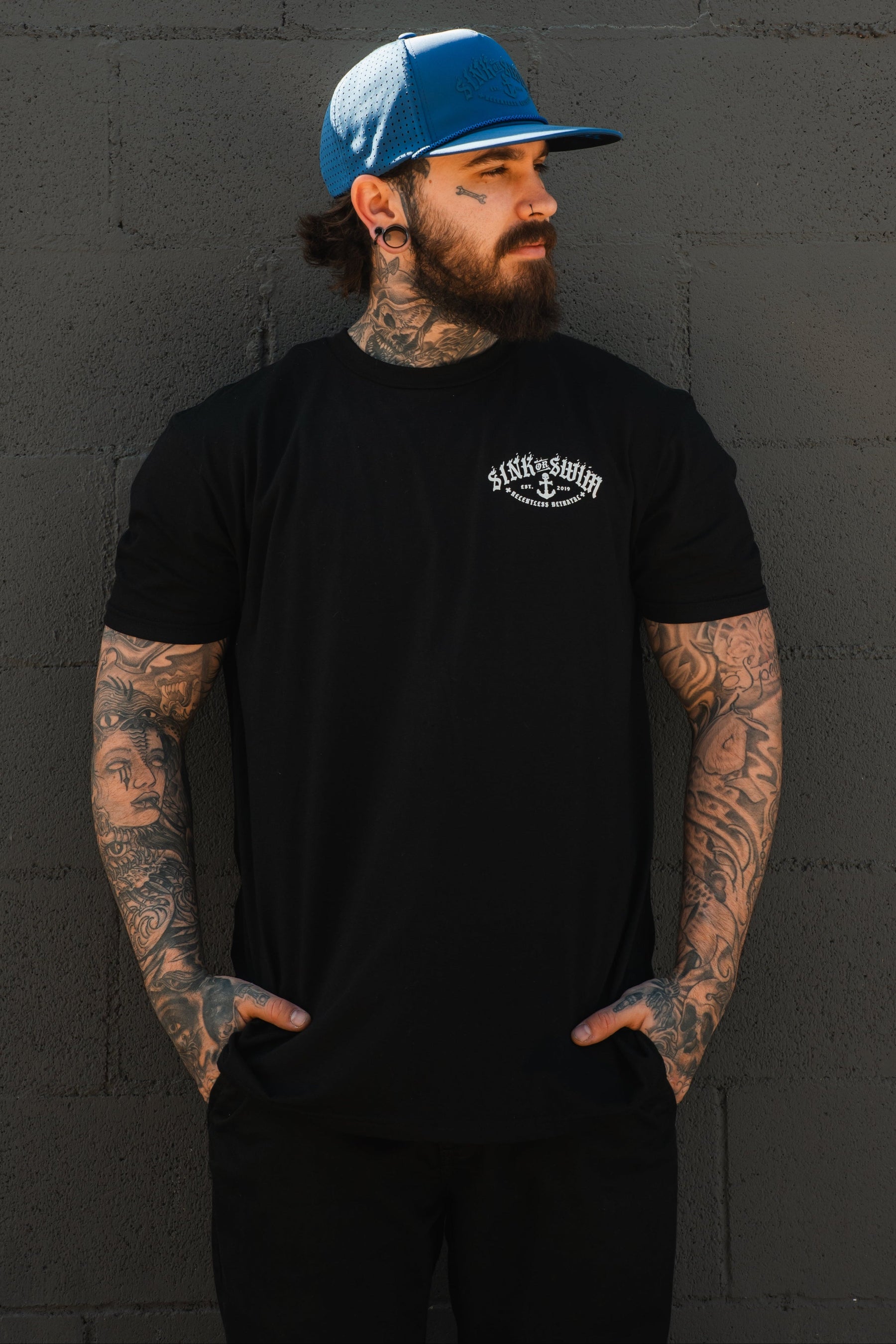 A bearded, tattooed man stands against a gray brick wall, wearing a Relentless Betrayal Dark Waters SOS T-Shirt, black pants, and a blue baseball cap. He looks to the side with his hands in his pockets.